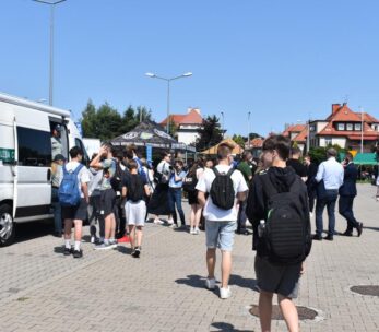 Grupa ludzi, głównie młodych, gromadzi się w pobliżu białej furgonetki i namiotów na imprezie plenerowej w słoneczny dzień. Niektórzy stoją w kolejce, inni spacerują. Wokół znajdują się budynki z czerwonymi dachami, zielone drzewa i czyste, błękitne niebo.