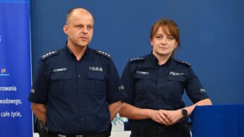 Dwoje policjantów, mężczyzna i kobieta, stoją obok siebie w ciemnoniebieskich mundurach z odznakami Policja. Wyglądają poważnie, zwróceni przodem do niebieskiej ściany z częściowo widocznym tekstem po lewej stronie i niebieskim podium po prawej stronie.