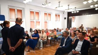 Grupa ludzi siedzi w rzędach krzeseł w jasnym pomieszczeniu, uważnie patrząc na dwóch funkcjonariuszy policji, którzy stoją z przodu. Funkcjonariusze przemawiają do publiczności. Pokój ma drewniane podłogi, białe ściany i okna z pomarańczowymi żaluzjami.