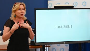 Kobieta o blond włosach gestykuluje serdecznie, kładąc ręce na klatce piersiowej, stojąc obok dużego ekranu dotykowego wyświetlającego polskie słowa UTUL SIEBIE. Wygląda na to, że jest w trakcie przemowy w klasie lub na seminarium.