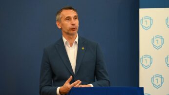 Mężczyzna w niebieskim garniturze przemawia na podium przykrytym niebieską tkaniną, gestykulując prawą ręką. Za nim znajduje się niebieska ściana i baner z logo niebieskiej tarczy. Po lewej stronie na stole widoczne są ulotki lub broszury.