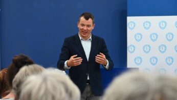 Mężczyzna w granatowej marynarce i jasnej koszuli gestykuluje podczas przemówienia do publiczności. Stoi przed niebieską ścianą i wyświetlaczem z powtarzającym się logo biało-niebieskiej tarczy. Kilka osób z siwymi włosami siedzi naprzeciwko niego.