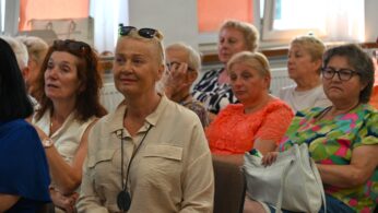 Grupa starszych kobiet siedzi blisko siebie w dobrze oświetlonym pokoju z pomarańczowymi zasłonami i dużymi oknami, uważnie obserwując coś poza kamerą. Większość jest ubrana swobodnie; jedna kobieta z przodu nosi okulary przeciwsłoneczne na głowie i beżową koszulę, podczas gdy inna ma na sobie jasnopomarańczową koronkę.
