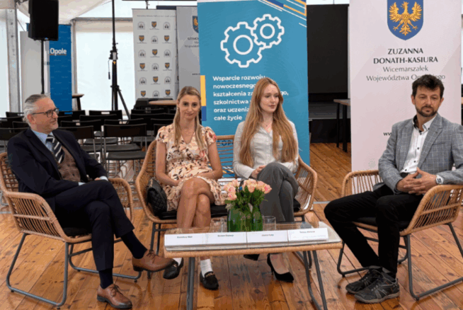 Cztery osoby siedzą na wiklinowych krzesłach wokół szklanego stołu z kwiatami, w namiocie eventowym z drewnianą podłogą. Za nimi znajdują się banery z polskim tekstem, logo i herbem. Dwóch mężczyzn ubranych jest w garnitury, a dwie kobiety w jasne sukienki. Zwróceni są twarzami do przodu, wyglądając na uważnych.