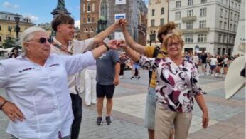 Cztery osoby - dwie starsze kobiety i dwóch młodszych mężczyzn - stoją trzymając się za ręce w kręgu, uśmiechając się na tętniącym życiem europejskim placu. Za nimi znajduje się gotycki kościół z rusztowaniami i ozdobnymi budynkami. Scenę dopełniają inni ludzie i błękitne niebo z chmurami.