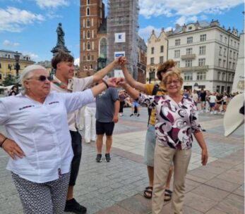 Cztery osoby - dwie starsze kobiety i dwóch młodszych mężczyzn - stoją trzymając się za ręce w kręgu, uśmiechając się na tętniącym życiem europejskim placu. Za nimi znajduje się gotycki kościół z rusztowaniami i ozdobnymi budynkami. Scenę dopełniają inni ludzie i błękitne niebo z chmurami.