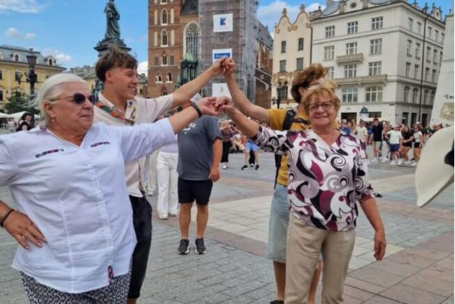 Cztery uśmiechnięte osoby, dwie starsze kobiety i dwóch młodych mężczyzn, trzymają się za ręce w kręgu, tańcząc na tętniącym życiem placu miejskim. W tle znajdują się zabytkowe budynki i wysoka wieża kościelna w remoncie, a nad nimi tłumy i częściowo zachmurzone niebo.