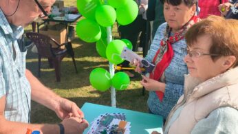 Mężczyzna przygotowuje balony na zewnętrznym stoisku z zielonymi balonami przywiązanymi do stojaka. Dwie starsze kobiety przyglądają się i trzymają broszury. Na stole leżą ulotki i nadmuchane balony. Inni ludzie i namioty są widoczne w słonecznym otoczeniu parku.