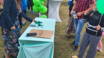 Ludzie stoją wokół stołu przykrytego jasnoniebieskim obrusem na imprezie plenerowej. Kobieta w kwiecistej spódnicy układa przedmioty na stole, podczas gdy inni, w tym dziecko trzymające zielony balon, patrzą na nią. W tle widoczne są zielone balony i namioty.