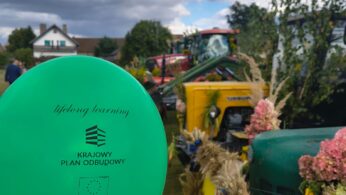 Ręka trzyma zielony balon z polskim tekstem o uczeniu się przez całe życie. W tle widać udekorowane zabytkowe traktory i kwiaty na zewnątrz pod częściowo zachmurzonym niebem, z widocznymi domami i drzewami. Scena sugeruje świąteczne wydarzenie społeczności.