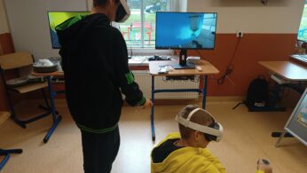 Dwóch chłopców w goglach VR bawi się wirtualną rzeczywistością w klasie; obraz z gogli wyświetlany jest na monitorze, widać proces lakierowania części karoserii samochodów.