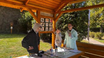 Kobieta z mikrofonem i dwoje dzieci (chłopcy) stoją pod drewnianą wiatą, przy stole informacyjnym, omawiając planszę z informacją o rozkładzie odpadów w czasie.