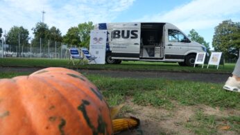 Mobilny autobus informacyjny (EduBus) z otwartymi drzwiami stoi na trawiastym terenie. Na pierwszym planie, rozmyta duża dynia, kolba kukurydzy i jarzębina.