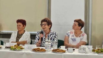 Trzy starsze kobiety siedzą przy stole nakrytym talerzami z jedzeniem, ciastkami, kubkami i butelkowaną wodą. Kobieta w centrum, trzymająca mikrofon, wydaje się mówić. Wszystkie trzy noszą okulary i swobodne ubrania, z neutralnymi ścianami w tle.