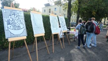 Cztery duże szkice/kolorowanki na sztalugach stoją w parku na zewnątrz. Na pierwszym planie grafika z owocami w misie. Kilku nastolatków stoi w tle, patrząc w kierunku stanowiska.