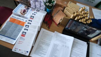 Stół pokryty jest broszurami, broszurami, długopisami, ołówkami i zwiniętymi papierami. Otwarty laptop wyświetla rozmowę wideo z kilkoma osobami. Stosy papieru i artykułów promocyjnych są uporządkowane, co sugeruje targi pracy lub wydarzenie informacyjne.