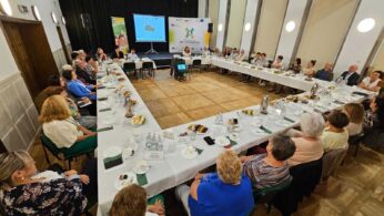 Duża grupa ludzi, w większości seniorów, siedzi wokół stołu w kształcie litery U z przekąskami i napojami w jasnej sali konferencyjnej. Z przodu prelegent prezentuje slajdy na ekranie. Przestrzeń zdobią banery i żyrandol.