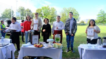 Grupa uczniów i dorosłych (kobieta) w ubraniach gastronomicznych (fartuchy, białe koszule) stoi za dwoma stołami z poczęstunkiem (napoje, owoce, ciasta) i kawą na zewnątrz. Dwoje uczniów trzyma tabliczki z napisami 