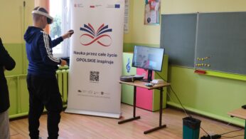 Nastolatek w granatowej bluzie stoi, używając gogli VR i kontrolerów, obok białego banera promocyjnego z napisem 