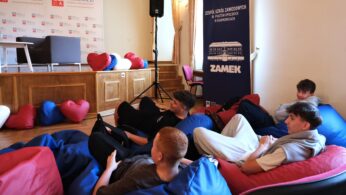 Czterech nastoletnich chłopców relaksuje się na kolorowych workach z fasolą w pokoju z drewnianą boazerią i sceną. Za nimi znajduje się duży niebieski baner z napisem 
