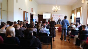 Grupa ludzi siedzi naprzeciwko ekranu projektora i prezenterów w jasnym pomieszczeniu z żyrandolami i dużymi oknami. Jedna kobieta stoi w przejściu, filmując lub fotografując wydarzenie. Publiczność wydaje się być uważna, a niektóre banery i plakaty są wyświetlane z przodu.