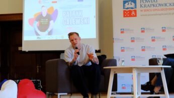 Mężczyzna siedzi na fotelu scenicznym i mówi do mikrofonu. Za nim znajduje się ekran, na którym wyświetlany jest Patryk Galewski z grafiką promocyjną. W tle znajdują się poduszki w kształcie serca, mały stolik ze szklankami wody oraz baner 