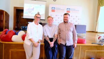 Trzy dorosłe osoby stoją razem w pomieszczeniu przed drewnianą sceną z banerami promocyjnymi i ekranem projekcyjnym za nimi. Dwóch mężczyzn i jedna kobieta pozują i uśmiechają się; w pobliżu rozrzucone są kolorowe worki z fasolą. Wygląda to na formalne wydarzenie lub prezentację.