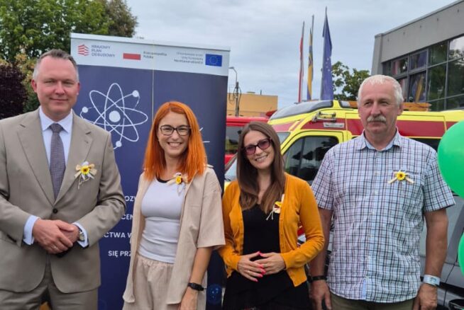 Na zdjęciu widać cztery osoby stojące na zewnątrz przy stoisku informacyjnym, na tle baneru z logotypami Unii Europejskiej i programu wsparcia. Osoby są uśmiechnięte, stoją obok stołu przykrytego zielonym obrusem, na którym znajdują się broszury, ulotki i gadżety promocyjne. W tle widać balony, samochód oraz budynek. Zdjęcie wykonano w ciągu dnia, w plenerze