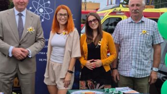 Na zdjęciu widać cztery osoby stojące na zewnątrz przy stoisku informacyjnym, na tle baneru z logotypami Unii Europejskiej i programu wsparcia. Osoby są uśmiechnięte, stoją obok stołu przykrytego zielonym obrusem, na którym znajdują się broszury, ulotki i gadżety promocyjne. W tle widać balony, samochód oraz budynek. Zdjęcie wykonano w ciągu dnia, w plenerze.