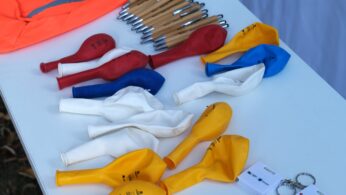 Nadmuchane balony w kolorach żółtym, białym, czerwonym i niebieskim leżą na stole obok kilku brązowych długopisów ze srebrnymi końcówkami, pomarańczowej torby oraz kilku białych plastikowych przedmiotów i breloczków.