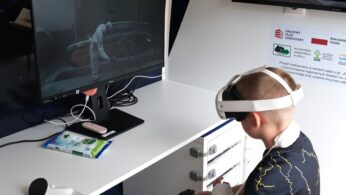 Dziecko noszące gogle VR siedzi przy biurku, używając kontrolerów do interakcji z wirtualną symulacją osoby pracującej przy samochodzie wyświetlaną na monitorze. Przestrzeń robocza obejmuje komputer, chusteczki czyszczące i plakaty instruktażowe na ścianie.