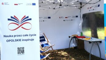 Biały namiot eventowy wyświetla duży pionowy baner z polskim tekstem, logo i kodem QR. Wewnątrz widoczne są składane krzesła, stół z broszurami, butelka, długopisy i zadaszona tablica. Namiot jest ustawiony na świeżym powietrzu na trawie z niewielką ilością światła słonecznego i cienia.