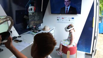 Młoda dziewczyna nosząca zestaw VR siedzi na czerwonym stołku w furgonetce technologicznej. Chłopiec trzyma w pobliżu inny zestaw słuchawkowy. Telewizor na ścianie wyświetla przemawiającego mężczyznę w garniturze. W przestrzeni znajdują się komputery, dokumenty i cyfrowy wystrój z kreskówkową pandą.