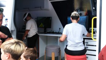 Wewnątrz mobilnego laboratorium naukowego kobieta siedzi przy biurku z komputerami, podczas gdy chłopiec nosi gogle VR w pobliżu. Dwóch chłopców o blond włosach i inne dziecko stoją na pierwszym planie, zaglądając do wnętrza furgonetki. Wewnątrz znajduje się sprzęt techniczny i stołki.