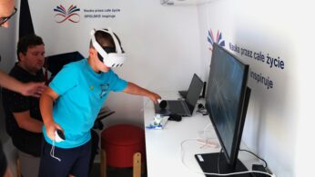 Chłopiec noszący gogle VR i trzymający kontrolery wchodzi w interakcję z monitorem komputera na białym biurku. Dwoje dorosłych siedzi w pobliżu. Na ścianie znajduje się polski tekst i logo. Otoczenie wygląda na edukacyjne, z widocznym sprzętem technologicznym i materiałami promocyjnymi.
