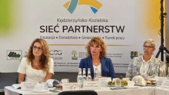 Trzy kobiety siedzą przy stole konferencyjnym z tabliczkami z nazwiskami, mikrofonami i butelkami wody. Za nimi znajduje się baner z napisem Sieć Partnerstw z kolorową grafiką i logo organizacji. Otoczenie wydaje się formalne i biznesowe.