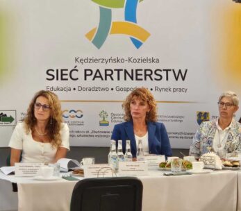 Trzy kobiety siedzą przy stole konferencyjnym z tabliczkami z nazwiskami, mikrofonami i butelkami wody. Za nimi znajduje się baner z napisem Sieć Partnerstw z kolorową grafiką i logo organizacji. Otoczenie wydaje się formalne i biznesowe.