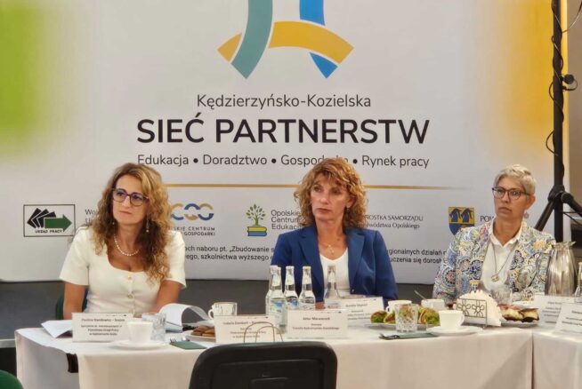 Trzy kobiety siedzą przy stole konferencyjnym z tabliczkami z nazwiskami, mikrofonami i butelkami wody. Za nimi znajduje się baner z napisem Sieć Partnerstw z kolorową grafiką i logo organizacji. Otoczenie wydaje się formalne i biznesowe.