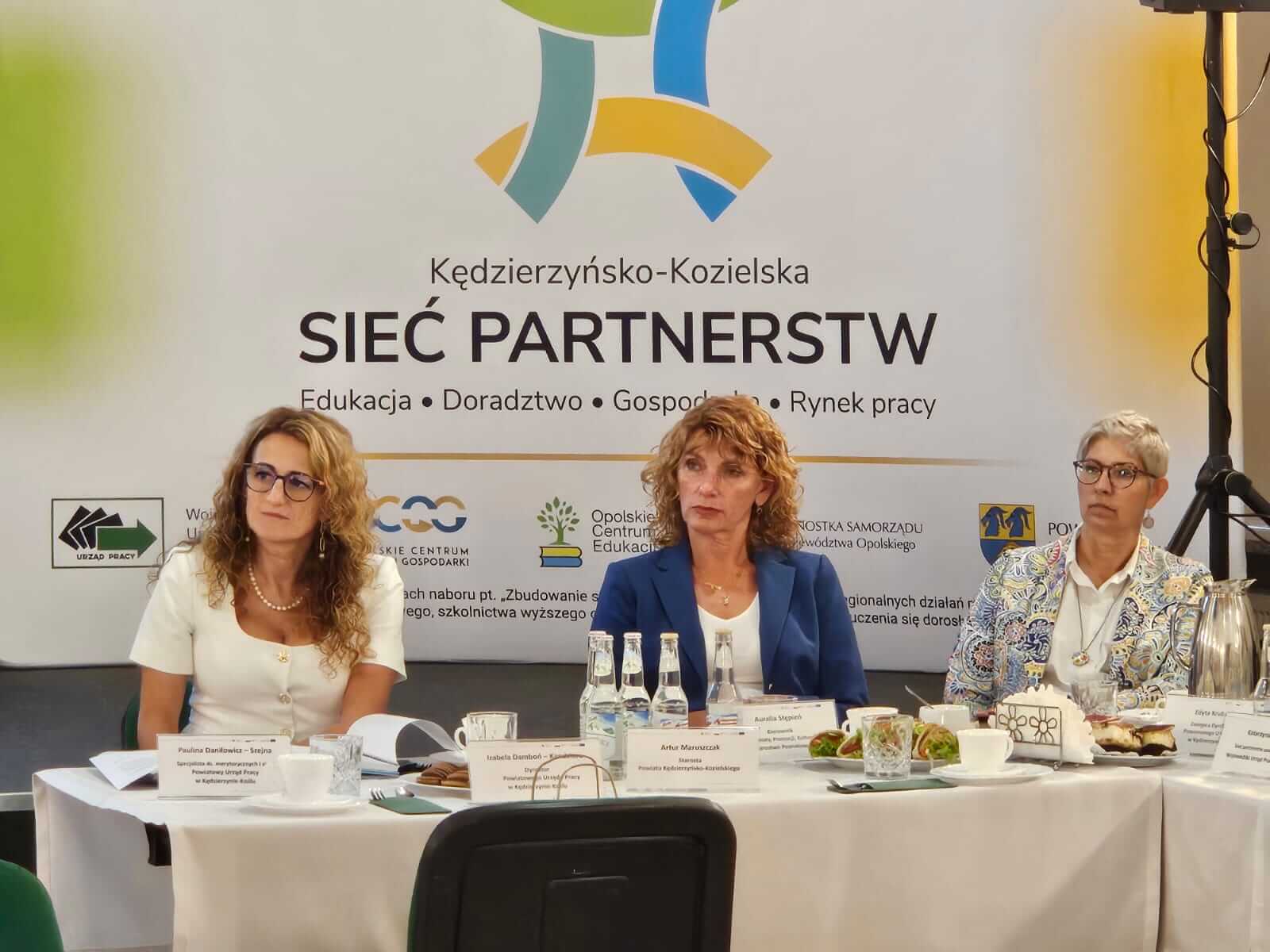 Trzy kobiety siedzą przy stole konferencyjnym z tabliczkami z nazwiskami, mikrofonami i butelkami wody. Za nimi znajduje się baner z napisem Sieć Partnerstw z kolorową grafiką i logo organizacji. Otoczenie wydaje się formalne i biznesowe.