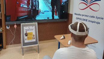 Osoba siedząca tyłem w białej koszulce i goglach VR, obsługująca kontrolery, patrząca na duży ekran wyświetlający scenę z mechanikiem przy ciężarówce. W tle widać biały baner z napisem 