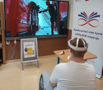 Osoba siedząca tyłem w białej koszulce i goglach VR, obsługująca kontrolery, patrząca na duży ekran wyświetlający scenę z mechanikiem przy ciężarówce. W tle widać biały baner z napisem "Nauka przez całe życie OPOLSKIE inspiruje".