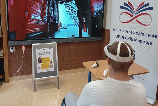 Osoba siedząca tyłem w białej koszulce i goglach VR, obsługująca kontrolery, patrząca na duży ekran wyświetlający scenę z mechanikiem przy ciężarówce. W tle widać biały baner z napisem "Nauka przez całe życie OPOLSKIE inspiruje".