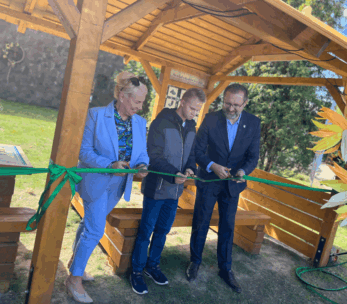 Trzy osoby – kobieta w jasnoniebieskim garniturze, młody chłopak w kurtce i mężczyzna w ciemnym garniturze – przecinają zieloną wstęgę nożyczkami pod drewnianą, zadaszoną konstrukcją na zewnątrz.