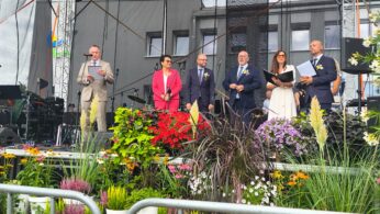 Sześć osób stoi na scenie z mikrofonami, ubranych formalnie. Przed sceną znajdują się kolorowe kwiaty i rośliny w białych doniczkach. Metalowa barykada oddziela publiczność od sceny. Światła sceniczne wiszą nad głową.