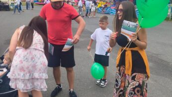 Mężczyzna w czerwonej koszuli i czapce stoi z kobietą we wzorzystej spódnicy trzymającą zielone balony i ulotki. W pobliżu stoi dziecko, również trzymające zielony balon. Inna kobieta w białej sukience pochyla się nad stołem na tętniącym życiem jarmarku z kolorowymi stoiskami.