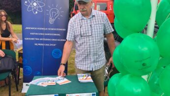 Mężczyzna w kraciastej koszuli i czapce stoi za zielonym stołem z broszurami i plakatami na imprezie plenerowej. W pobliżu znajdują się zielone balony i baner informacyjny dotyczący edukacji i innowacji. Drzewa i ludzie są widoczni w tle na trawiastym terenie.