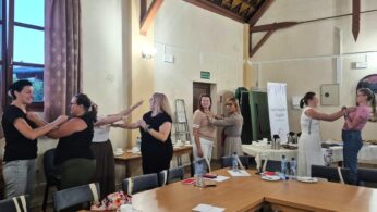 Siedem kobiet stoi w szeregu w pomieszczeniu, każda z nich uśmiecha się i delikatnie kładzie ręce na ramionach osoby stojącej przed nimi. Znajdują się w ciepło oświetlonym pomieszczeniu z wysokimi sufitami, stołami, butelkami wody, jedzeniem i flipchartem w tle.