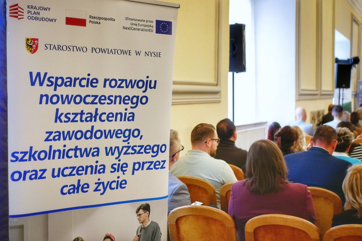 Baner informacyjny z napisem „wsparcie rozwoju kształcenia zawodowego i uczenia się przez całe życie. W tle uczestnicy siedzący tyłem na sali konferencyjnej.”