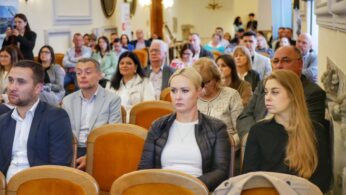 Uczestnicy konferencji siedzący na sali, uważnie słuchający prezentacji. Widoczne różne grupy wiekowe i formalna atmosfera wydarzenia.