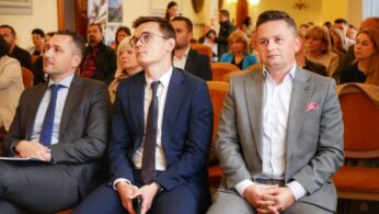 Trzech mężczyzn w garniturach siedzących w pierwszym rzędzie podczas konferencji. W tle pozostali uczestnicy, sala urzędowa.